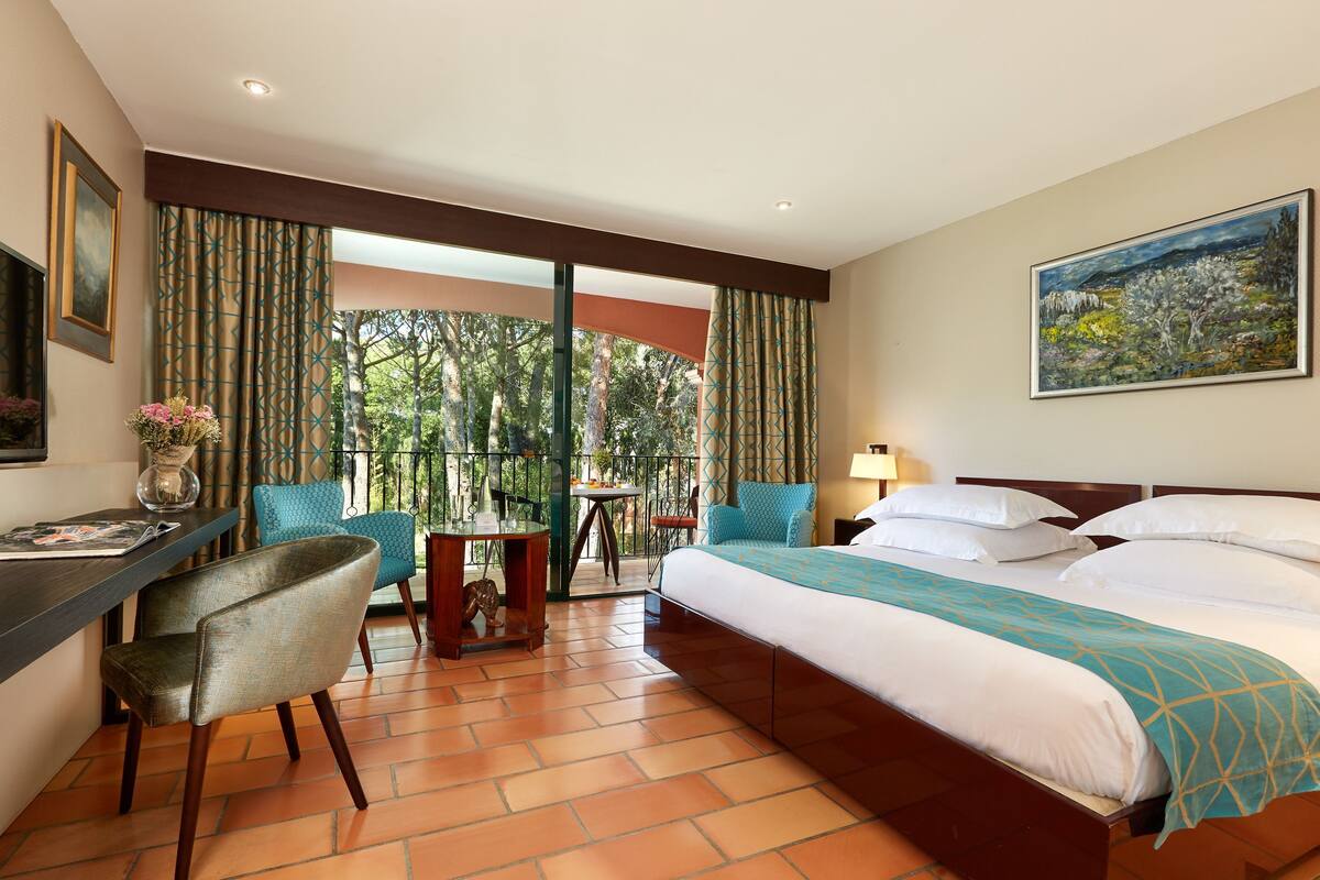 A hotel room with a bed, a desk, a chair, a balcony with a view of trees, and a painting on the wall.