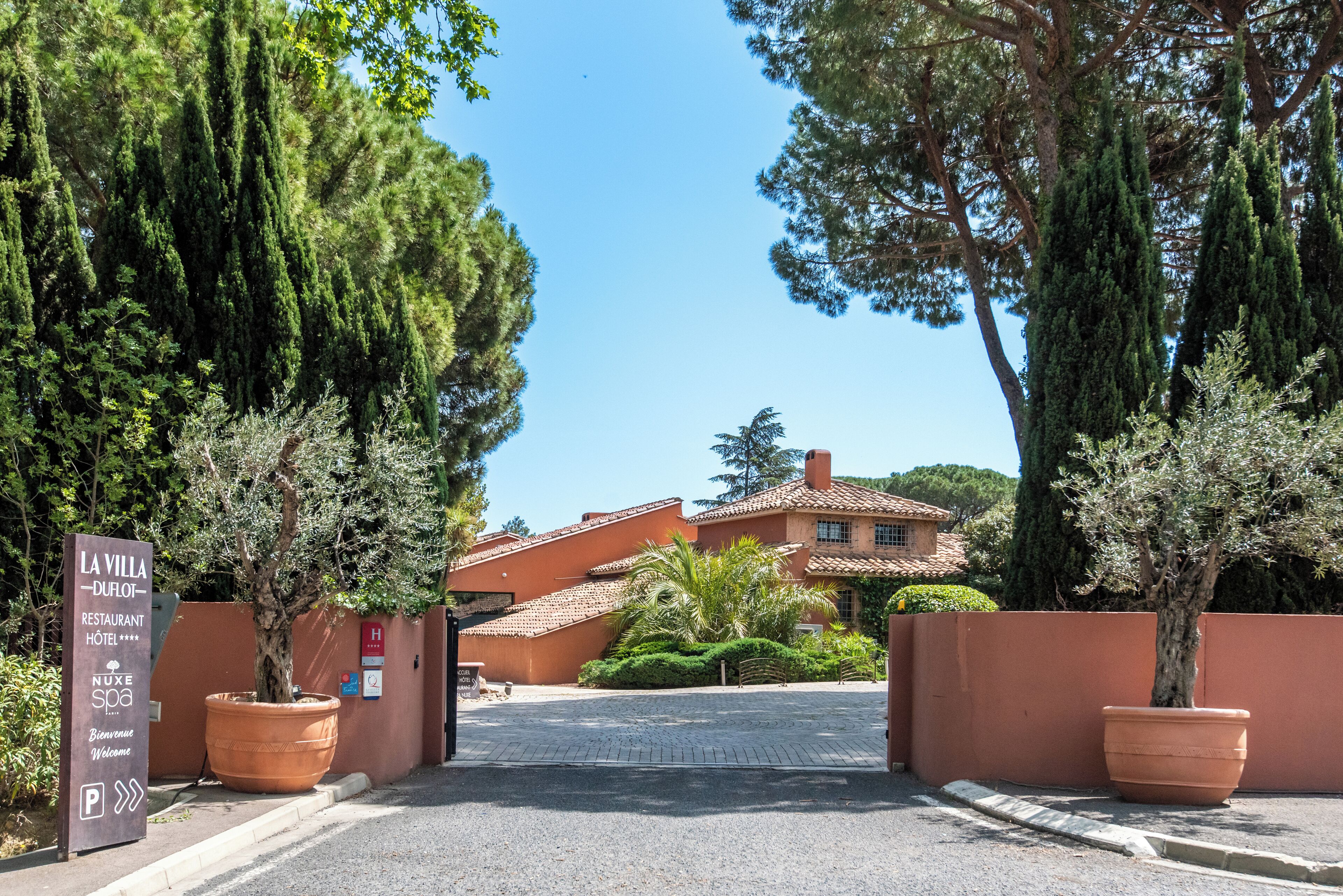 Una villa con acceso pavimentado, un letrero y un gran árbol en maceta.