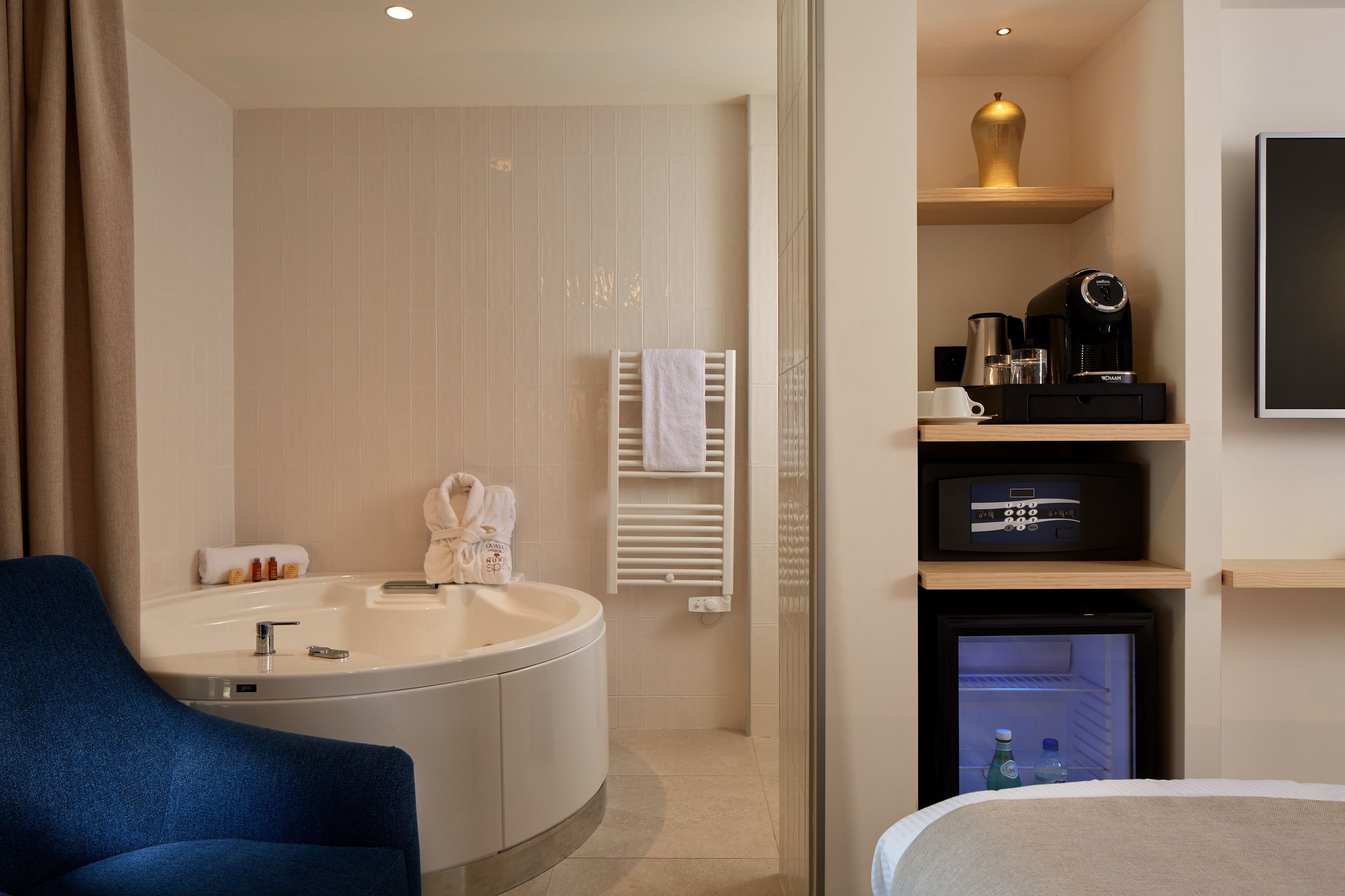 A modern hotel bathroom with a bathtub, a sink, a towel rack, a coffee machine, and a mini-fridge.