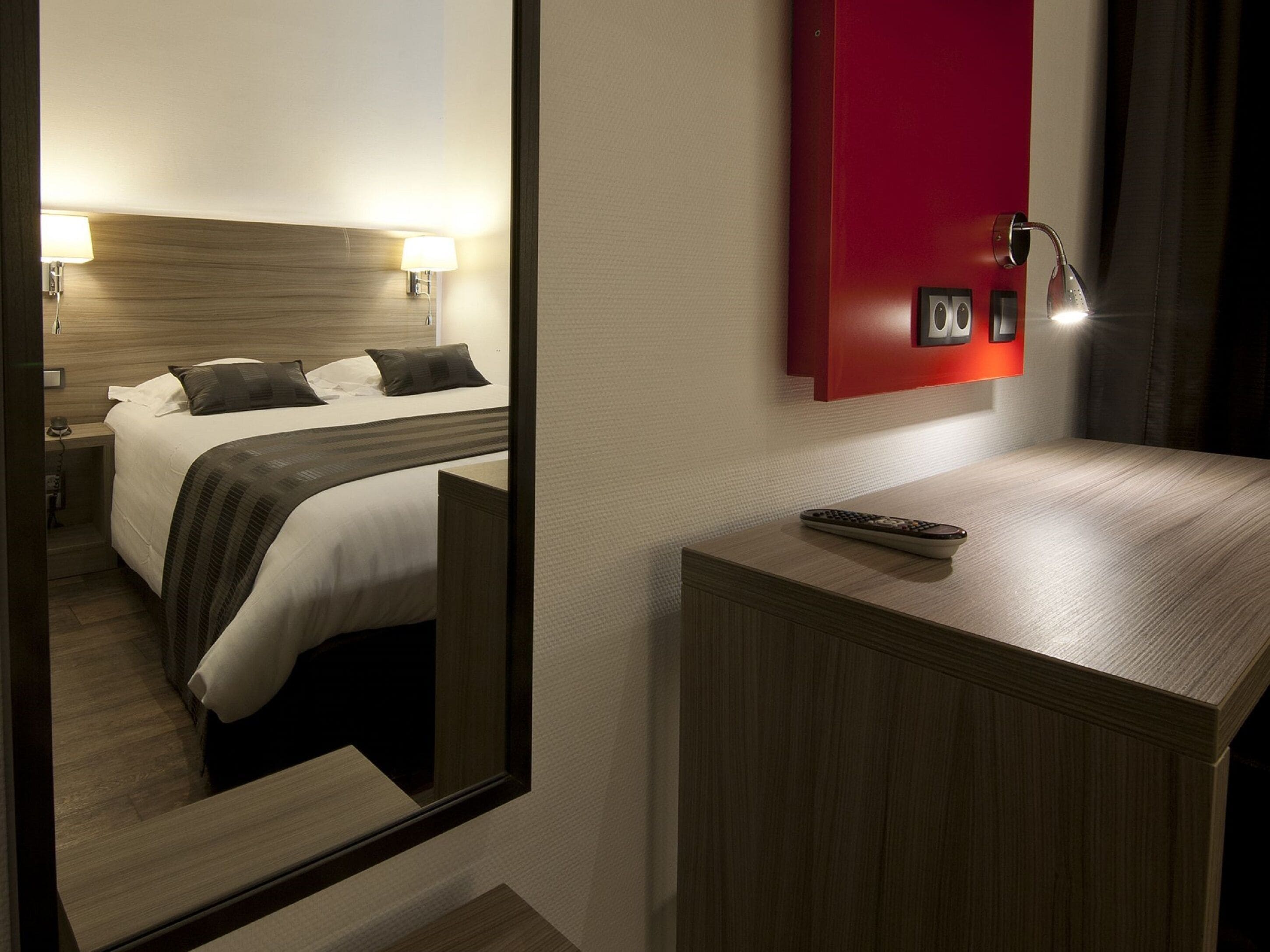 Habitación de hotel con cama, lámparas de noche, un espejo, una pared de acento roja y una mesita de noche con control remoto.