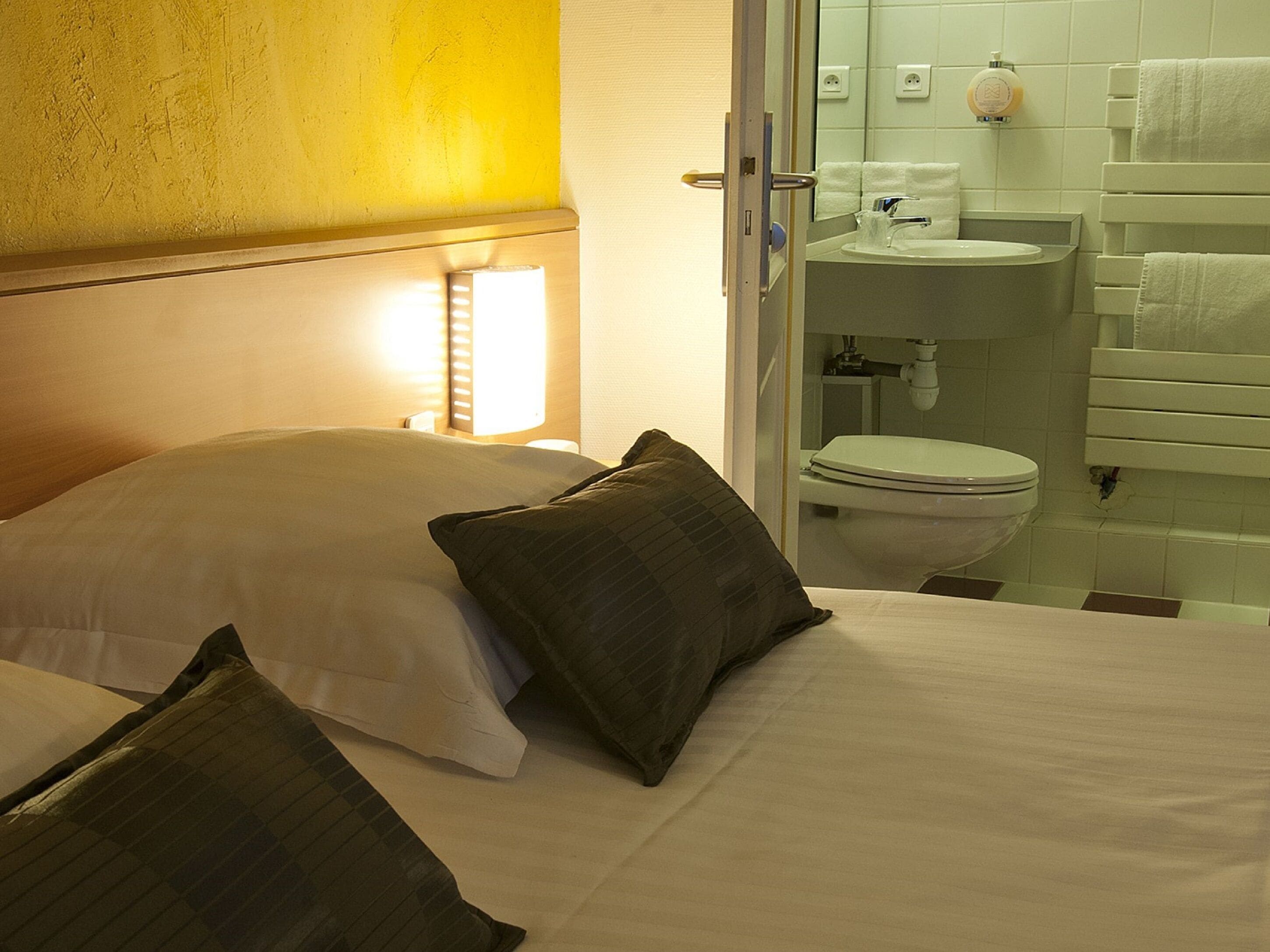 Une chambre d’hôtel comprenant un lit, des oreillers, une lampe de chevet et une salle de bain avec des toilettes, un lavabo et un porte-serviettes.