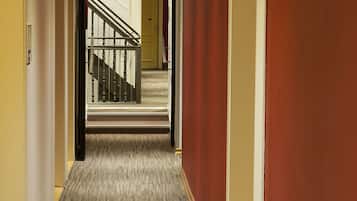 A hotel hallway with carpeted flooring, a staircase with wooden railings, and a door at the end.