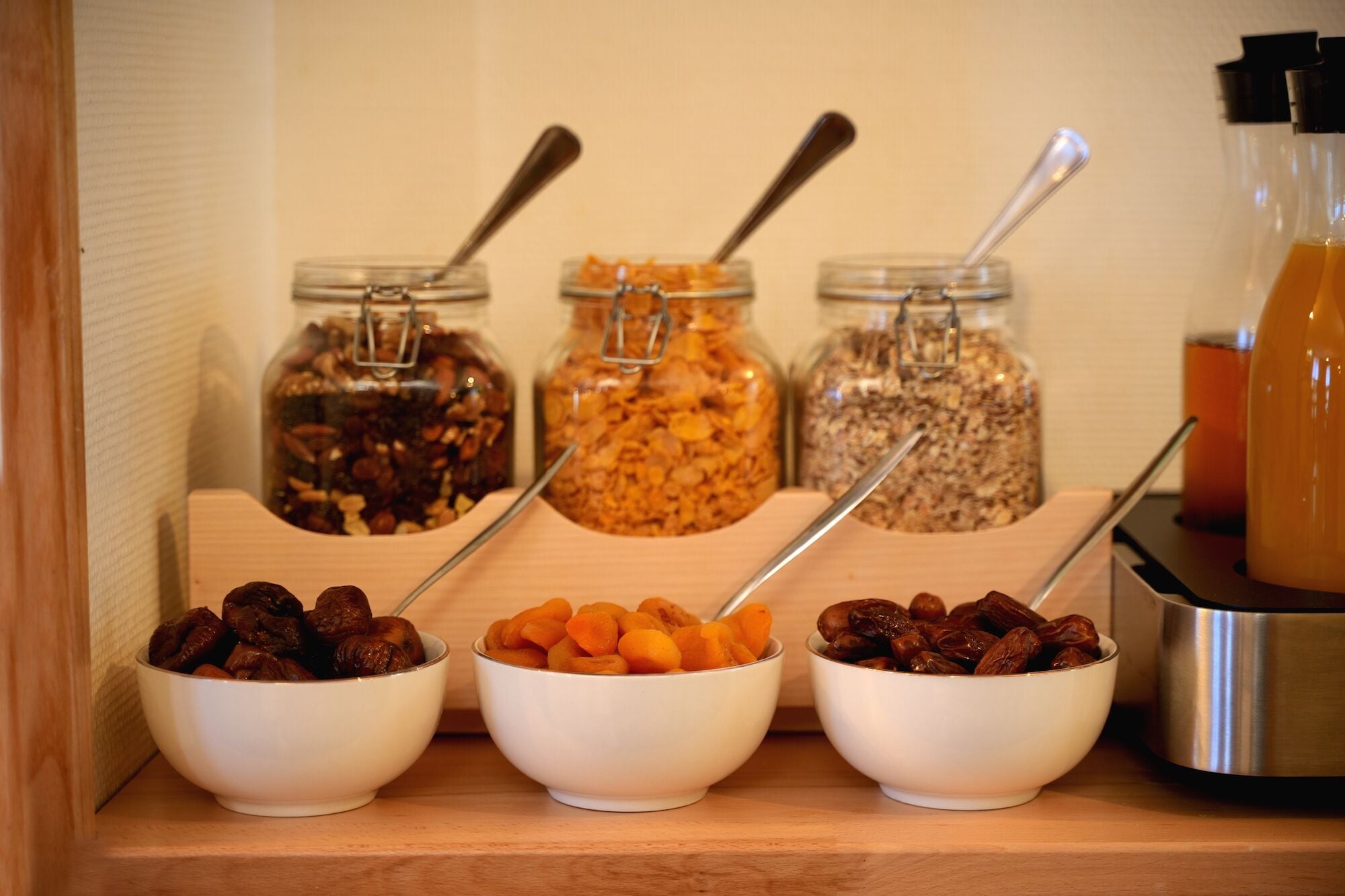 Three bowls of nuts and dried fruits on a wooden shelf, with jars of granola and a bottle of juice in the background.