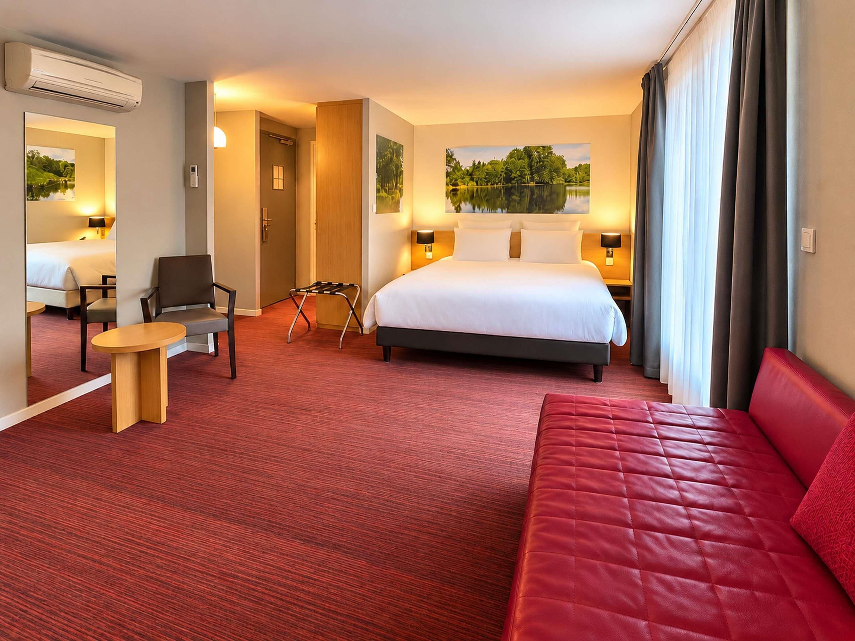 Une chambre d’hôtel avec un lit, un canapé rouge, une petite table, une chaise et un miroir.