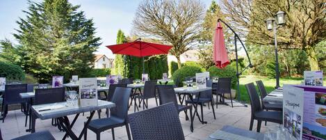 An outdoor dining area with tables and chairs, surrounded by trees and a clear sky.
