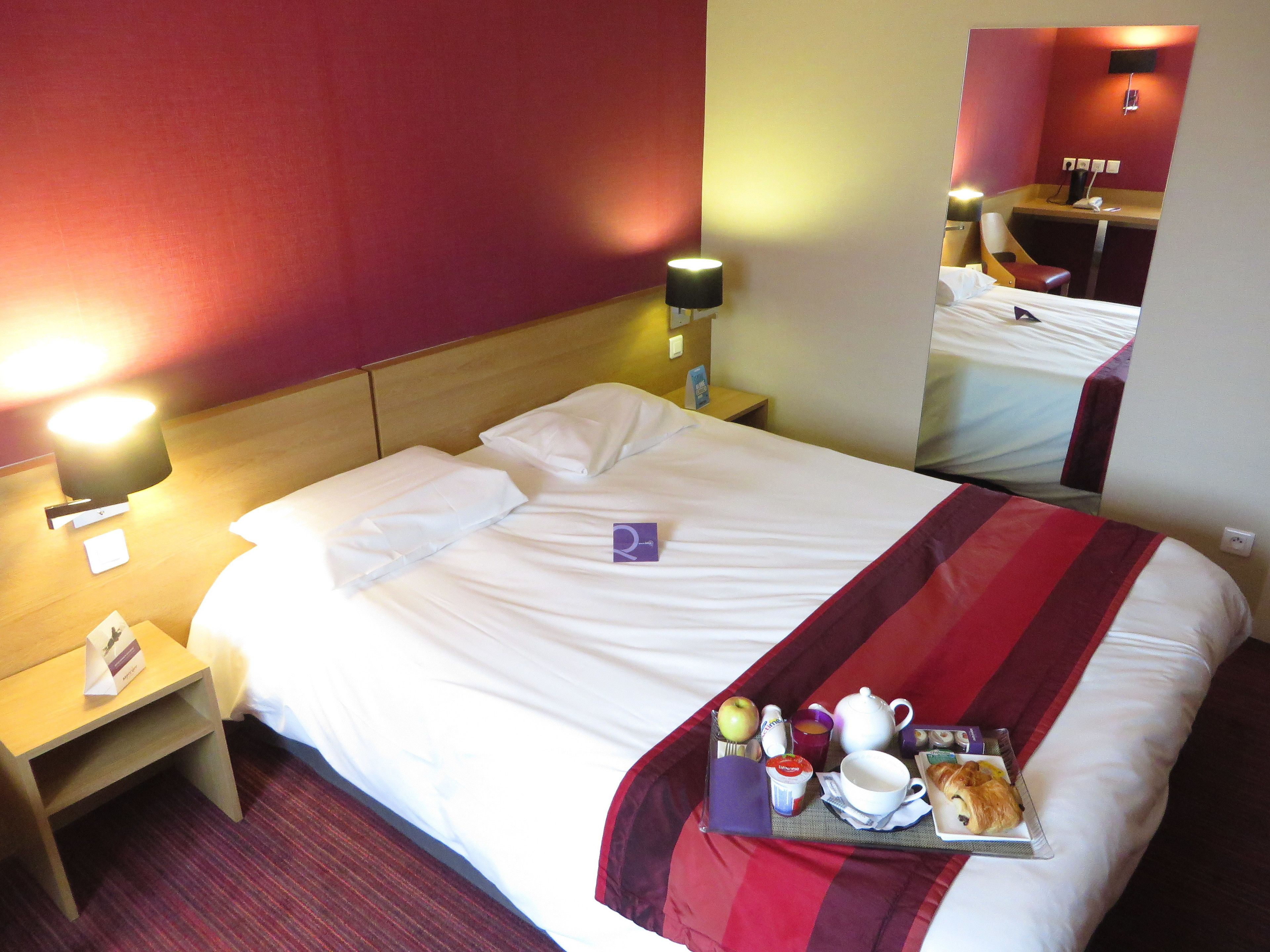 Une chambre d’hôtel avec un lit bien fait, une table de chevet avec une lampe et un plateau contenant des articles pour le petit-déjeuner.