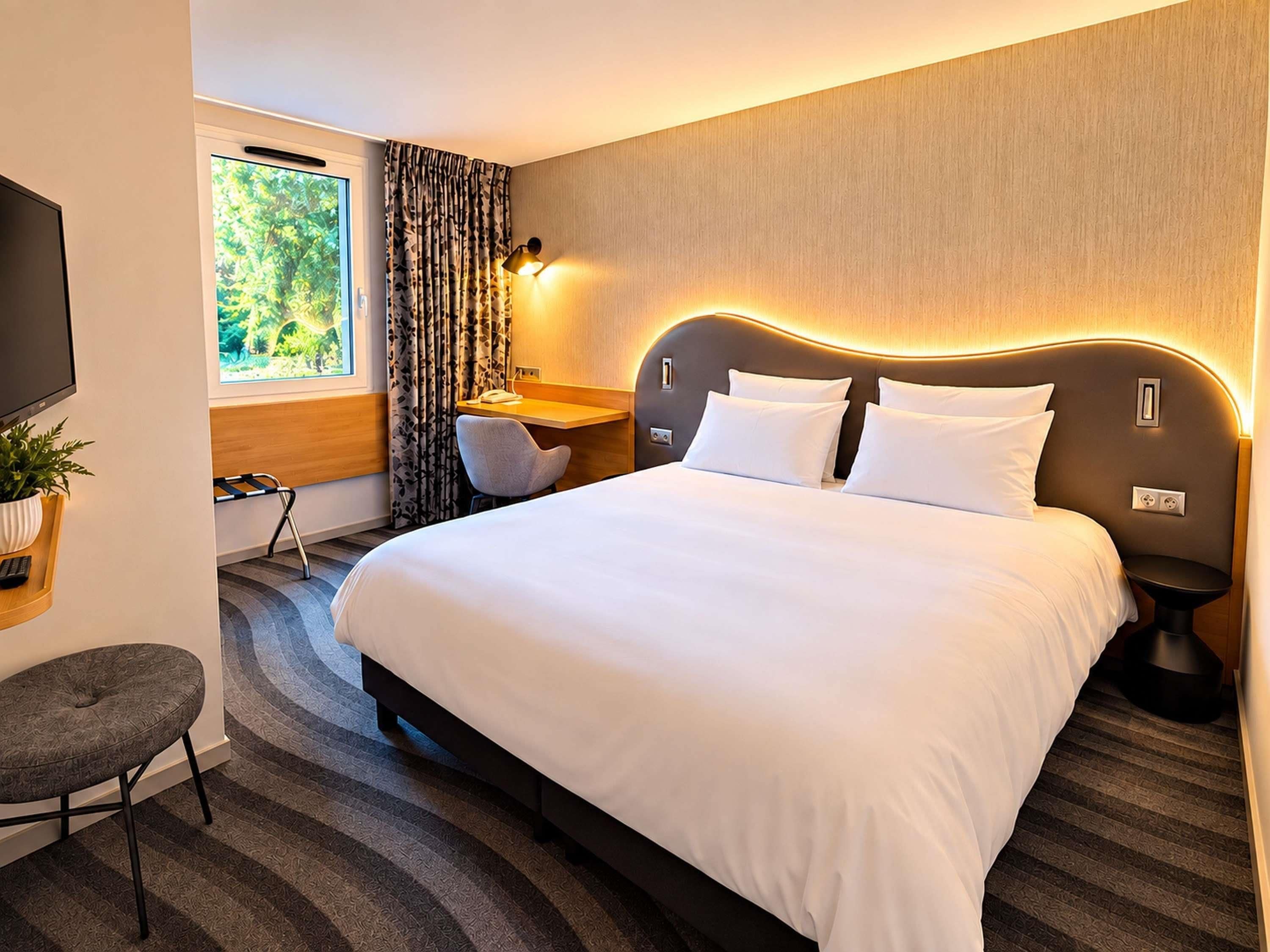 A hotel room with a large bed, a desk, a chair, a TV, and a window with curtains.
