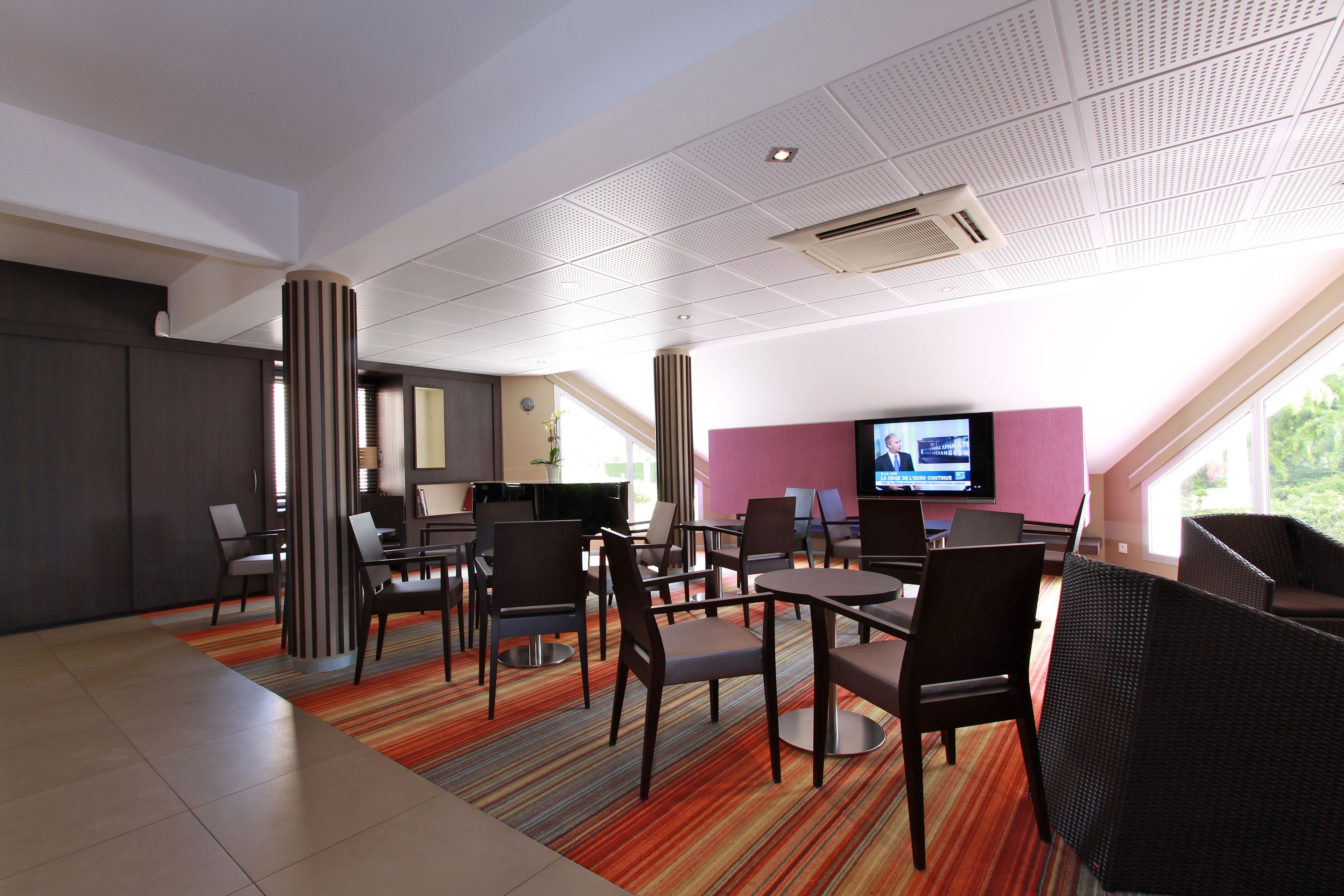 A modern hotel lobby with a mix of seating areas, including tables and chairs, a large flat-screen TV, and a large window offering a view of greenery outside.