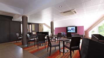 A modern hotel lobby with a mix of seating areas, including tables and chairs, a large flat-screen TV, and a large window offering a view of greenery outside.