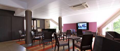 A modern hotel lobby with a mix of seating areas, including tables and chairs, a large flat-screen TV, and a large window offering a view of greenery outside.