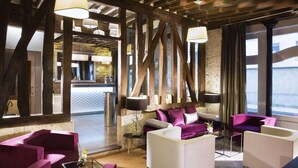 Un hall d’hôtel moderne avec des poutres en bois apparentes, un coin salon doté de canapés violets et blancs, et une vue sur la mer grâce à de grandes fenêtres.