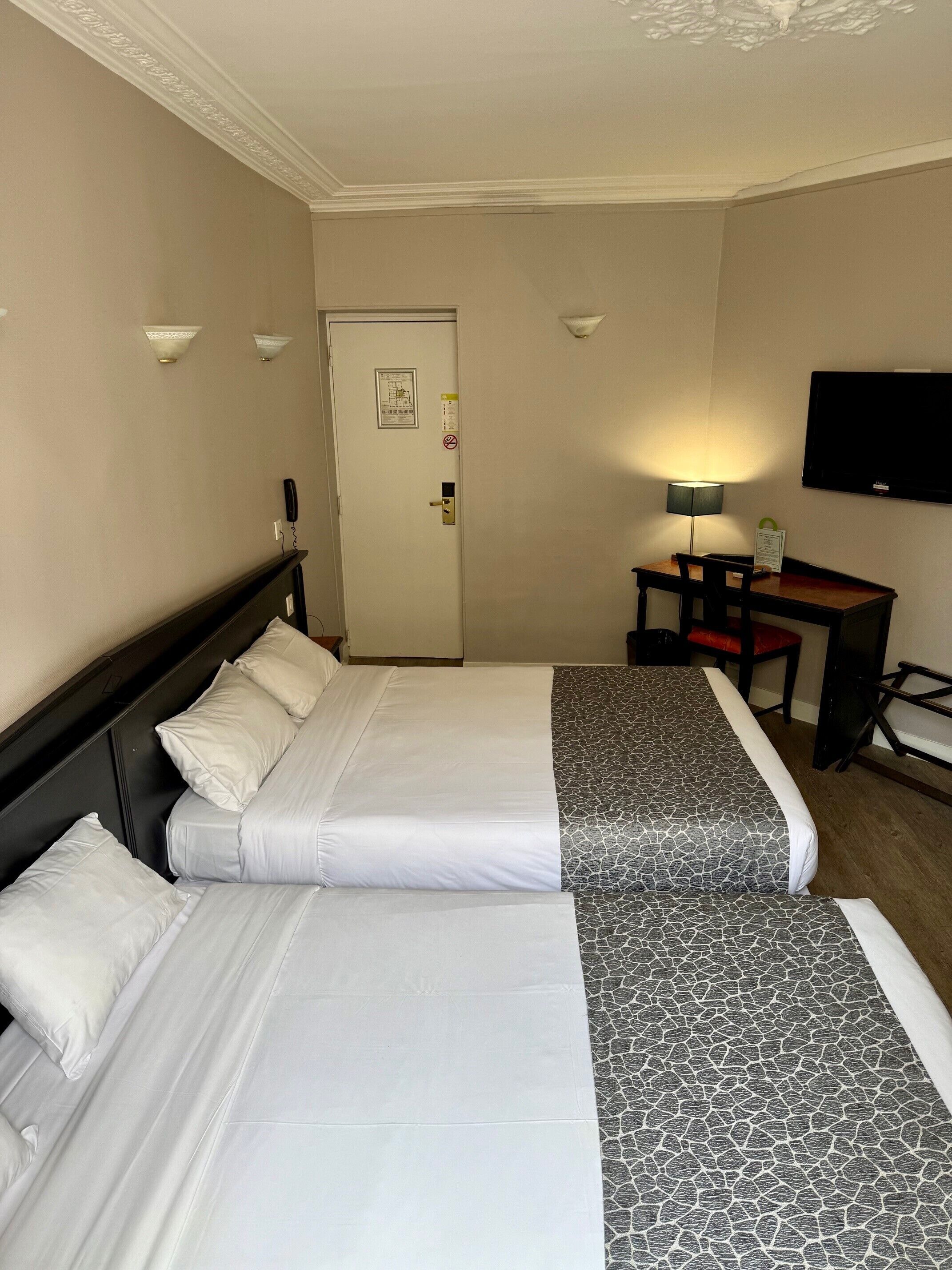 A hotel room with two beds, a desk, a chair, a TV, and a door with a sign.