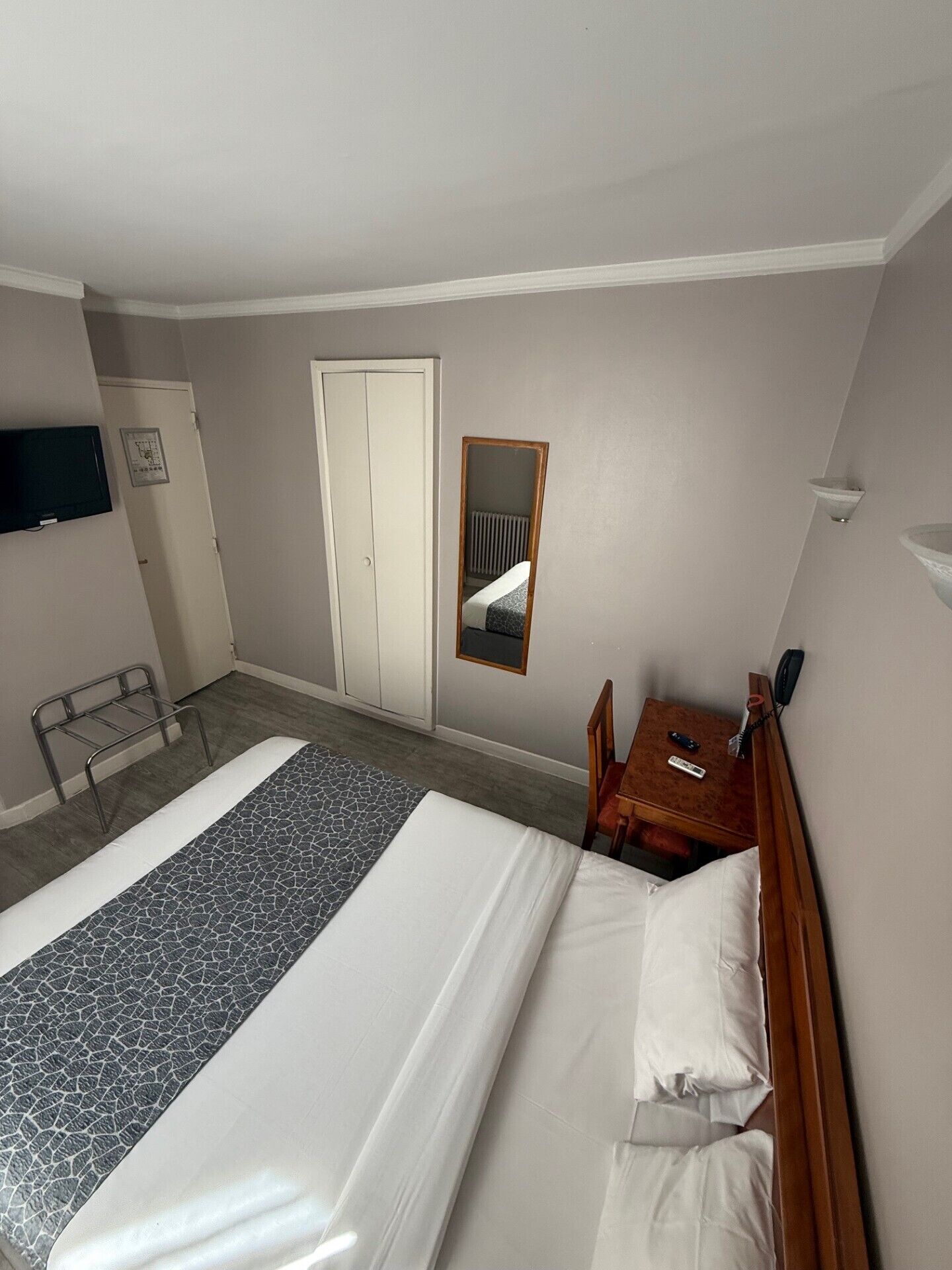 A hotel room with a bed, a wooden nightstand, a chair, a mirror, a radiator, and a television on the wall.