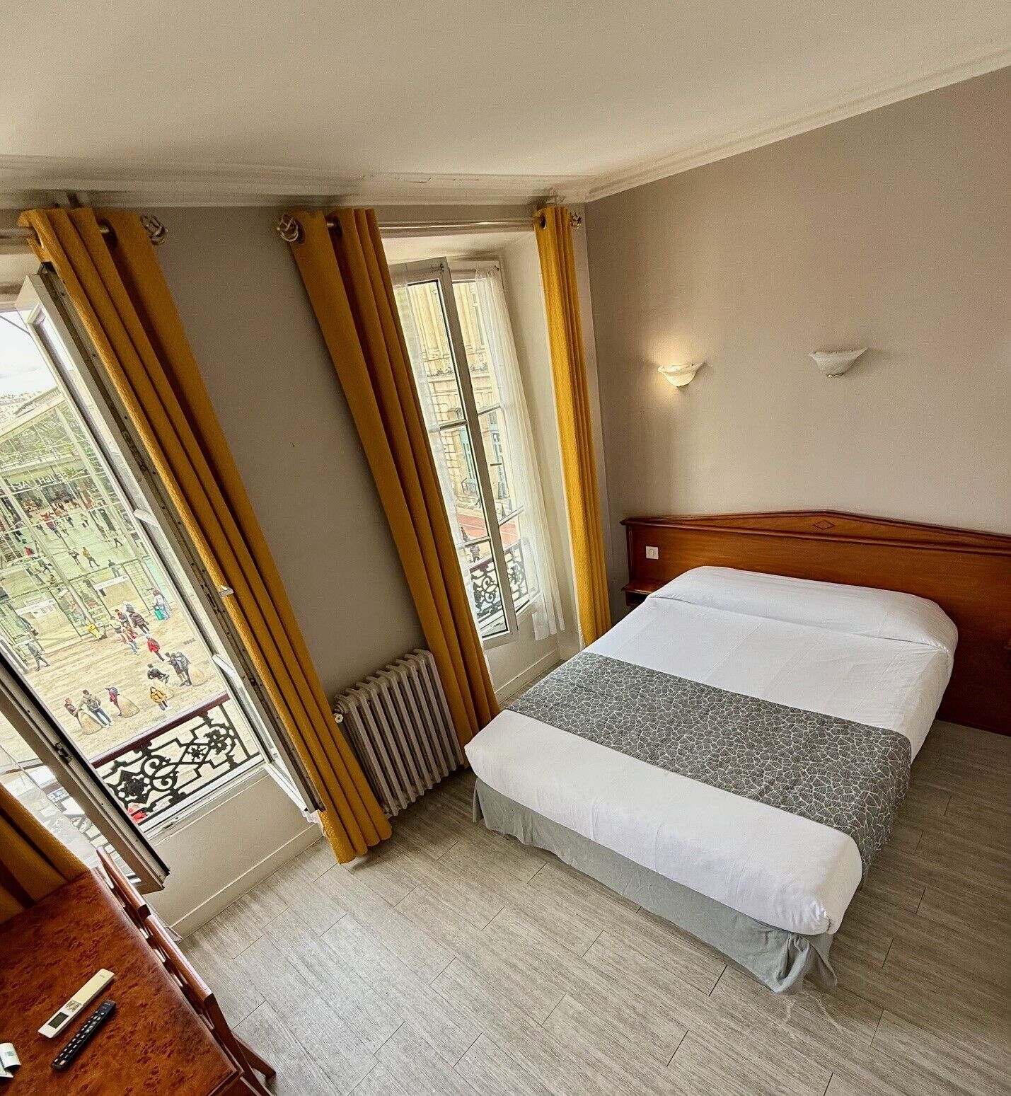 A hotel room with a bed, a wooden headboard, a window with curtains, a radiator, and a view of a plaza.
