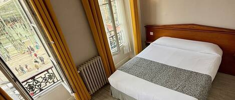 A hotel room with a bed, a wooden headboard, a window with curtains, a radiator, and a view of a plaza.