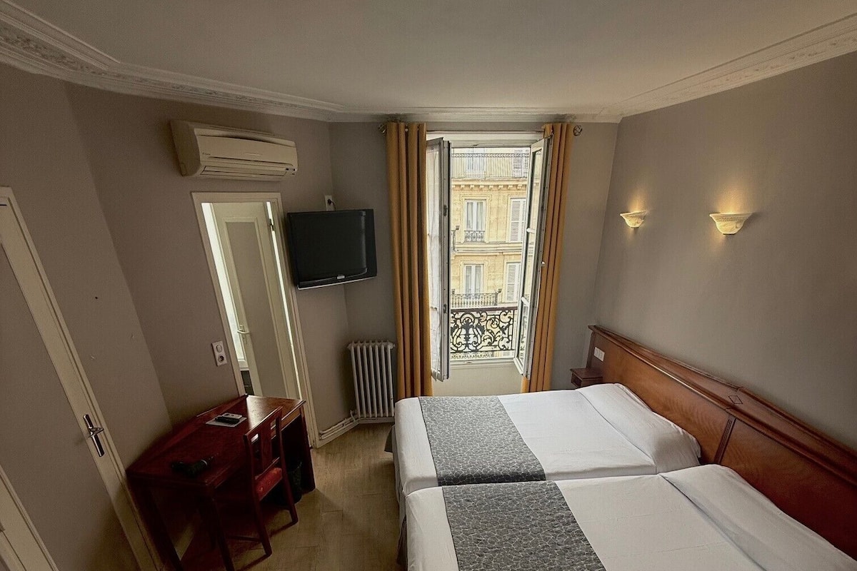 A hotel room with two beds, a desk, a TV, and a balcony with a view of buildings.