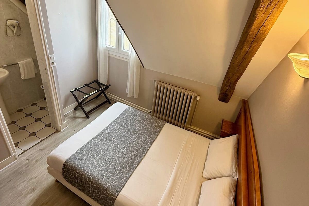 A bedroom with a sloped ceiling, a wooden headboard, a bed with white and gray bedding, a radiator, and a window with curtains.