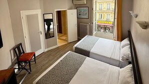 Una camera d'albergo con due letti, una sedia, una scrivania e vista su un edificio attraverso una porta aperta.