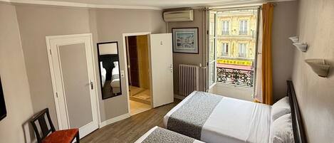 Habitación de hotel con dos camas, una silla, un escritorio y vista a un edificio desde una puerta abierta.