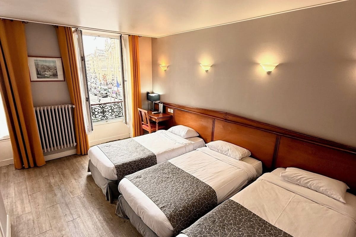 A hotel room with three beds, a wooden floor, a radiator, and a window with curtains.