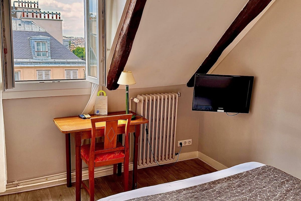 A bedroom with a bed, a desk with a chair, a radiator, a window with a view of buildings, and a TV mounted on the wall.