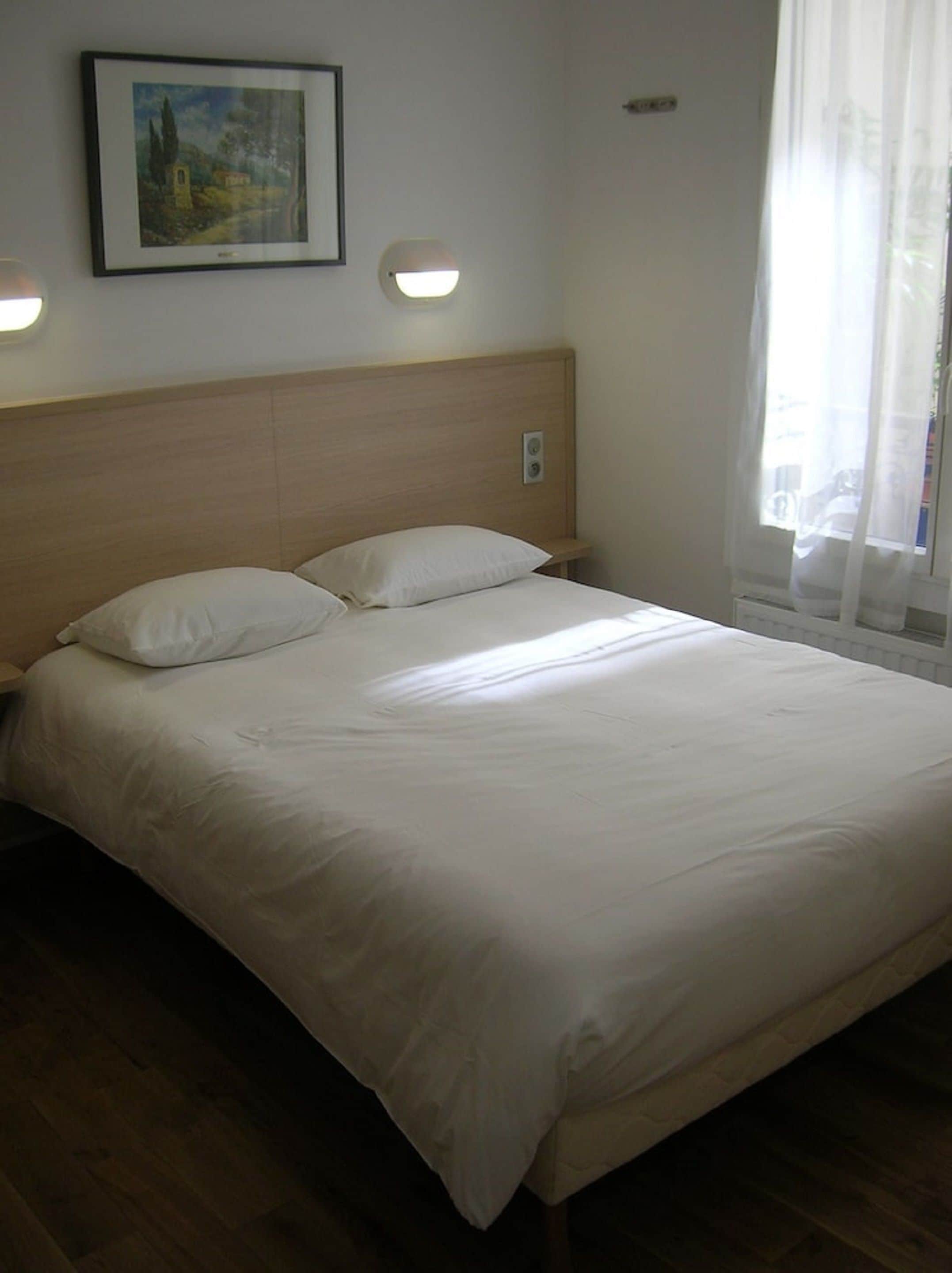 Un lit bien fait, avec du linge de lit blanc, dans une chambre dotée d’une tête de lit en bois, d’un tableau encadré au mur et d’une fenêtre ornée de rideaux légers.