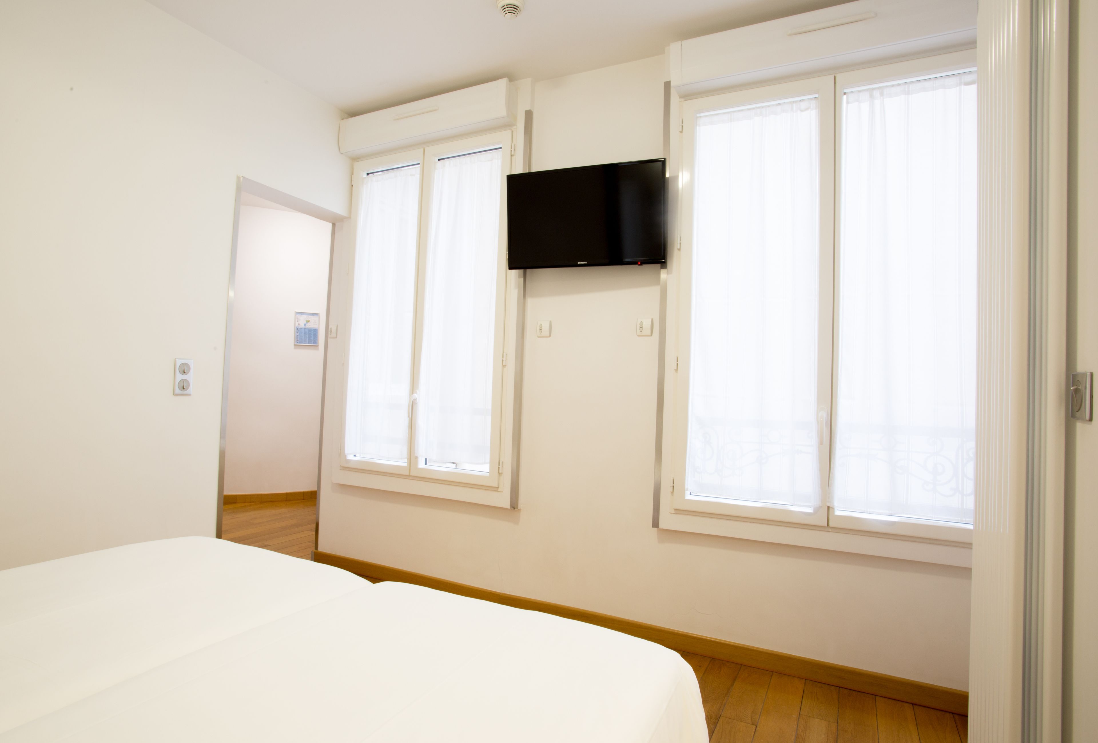 A hotel room with a bed, a television mounted on the wall, and two windows with curtains.