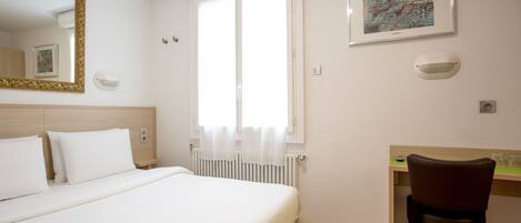 A hotel room with a bed, a desk, a chair, a window with curtains, a radiator, a mirror, and a framed picture on the wall.