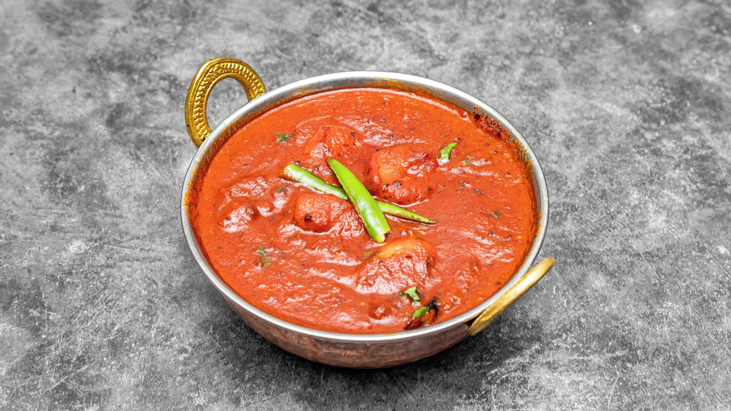 Un bol de curry rouge, agrémenté de piments verts et d’herbes aromatiques, est posé sur une surface grise texturée.