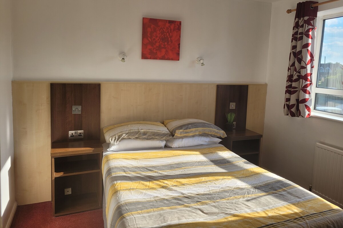 A bedroom with a bed, a nightstand, a shelf, a red artwork, and a window with curtains.