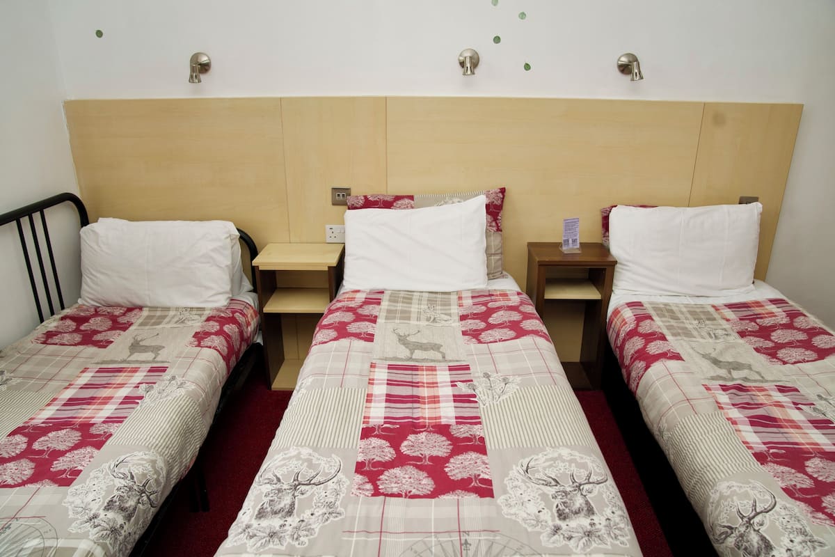 A hotel room with three single beds, each with a red and white patterned bedspread. There are wooden headboards and nightstands between the beds.