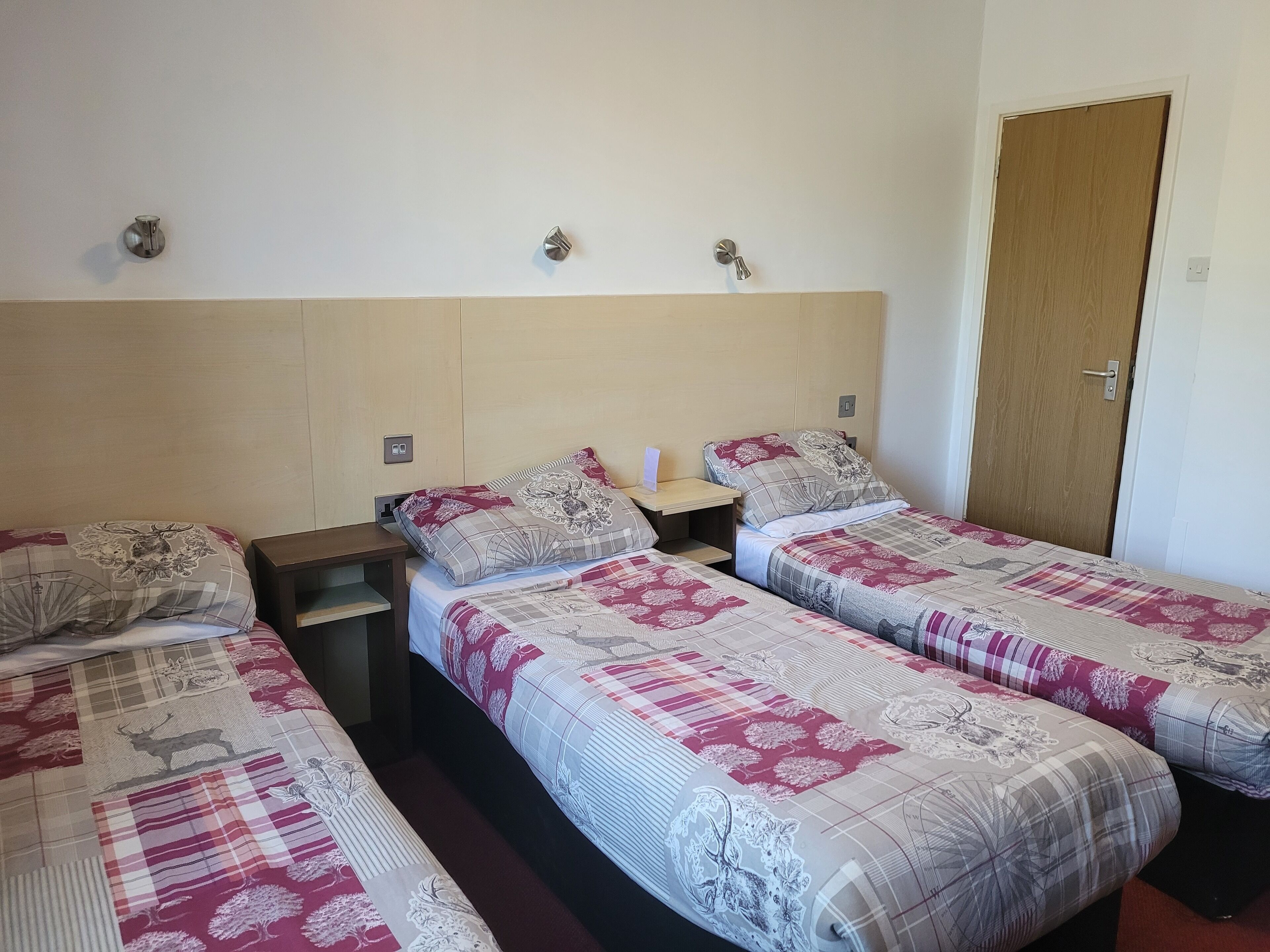Une chambre avec trois lits simples, chacun doté d’une couette à motifs, d’une table de chevet avec une lampe et d’une porte en bois.