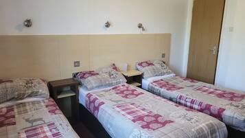 Une chambre avec trois lits simples, chacun doté d’une couette à motifs, d’une table de chevet avec une lampe et d’une porte en bois.