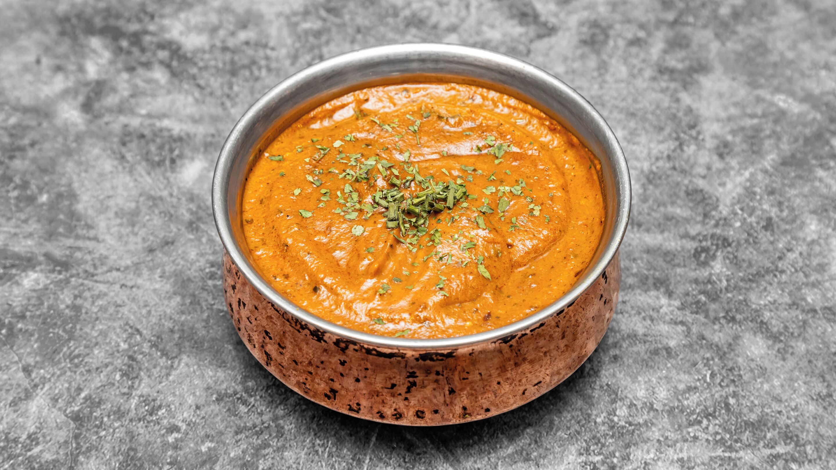 Een kom oranje curry met groene kruiden op een koperen bord.