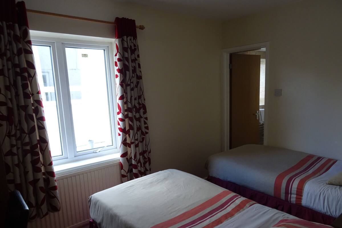 A room with two beds, a window with curtains, a radiator, and a door leading to another room.