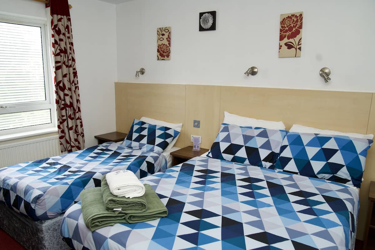 A hotel room with two beds, each with a geometric-patterned bedspread, a window with blinds, and a radiator.