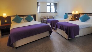 A hotel room with two beds, each with a purple bedspread and teal pillows, a small table with a chair, and a window with curtains. - Caladh Inn (Stornoway)