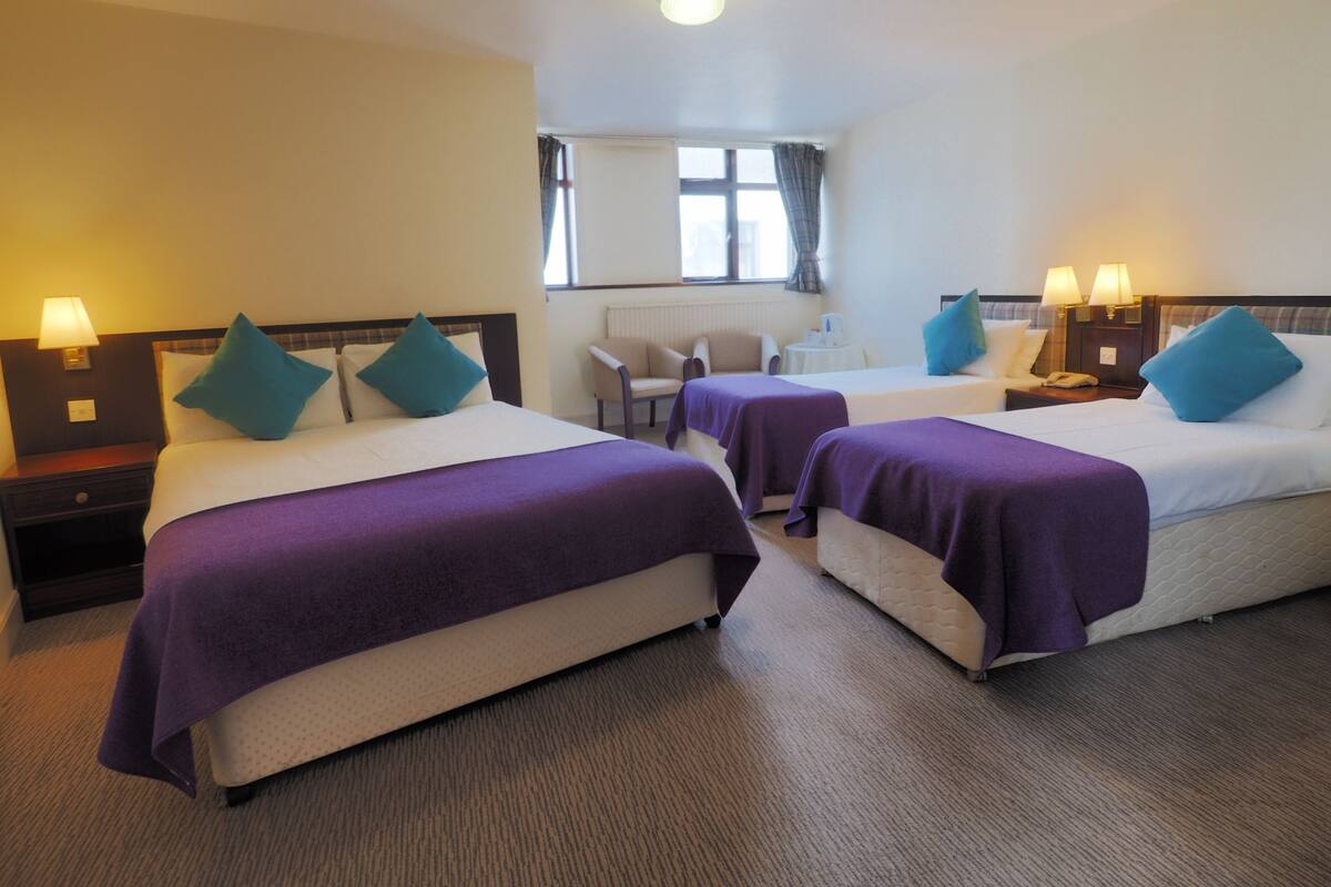 Habitación de hotel con dos camas, cada una con colcha púrpura y almohadas turquesa, una mesita con una silla y una ventana con cortinas.