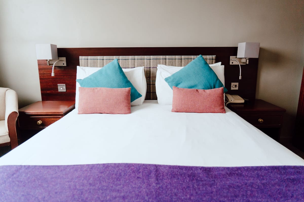 Una habitación de hotel con una cama grande, dos mesitas de noche, una silla y un cabecero con una lámpara.