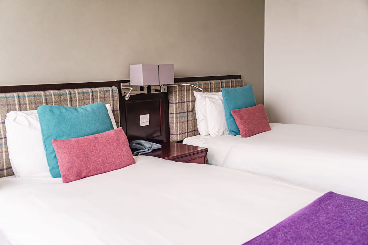 Habitación de hotel con dos camas, cada una con ropa blanca y almohadas coloridas. Hay una mesita de noche con una lámpara y un teléfono.
