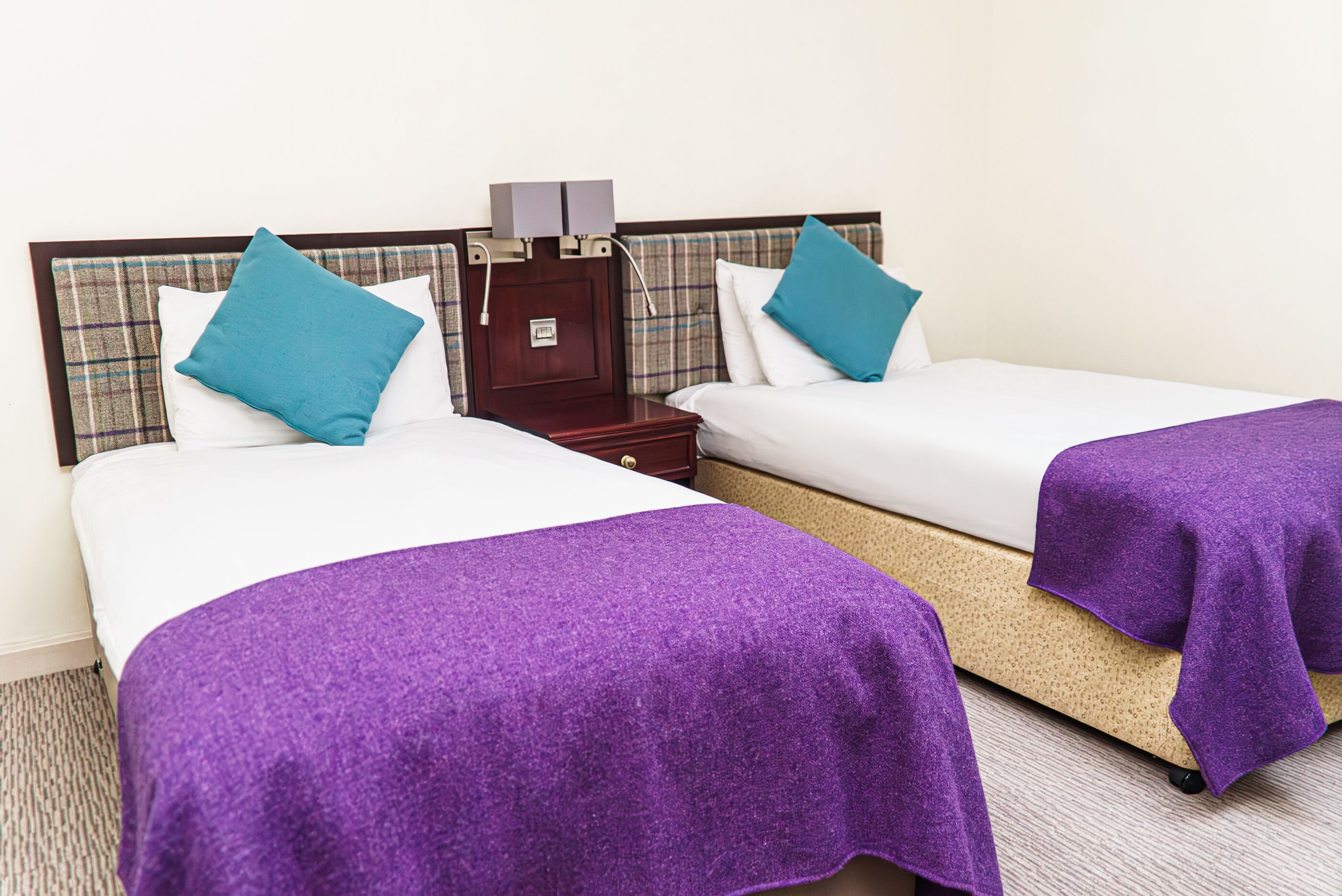 Habitación de hotel con dos camas individuales, cada una con un edredón morado y una almohada azul. Hay una mesita de noche de madera con una lámpara y una pequeña mesa entre las camas.