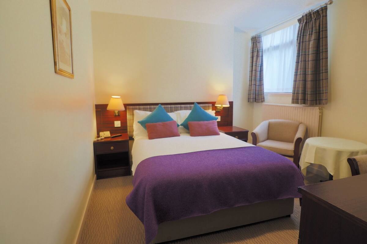 Una habitación de hotel con una cama, dos mesitas de noche con lámparas, una silla, una pequeña mesa y una ventana con cortinas.