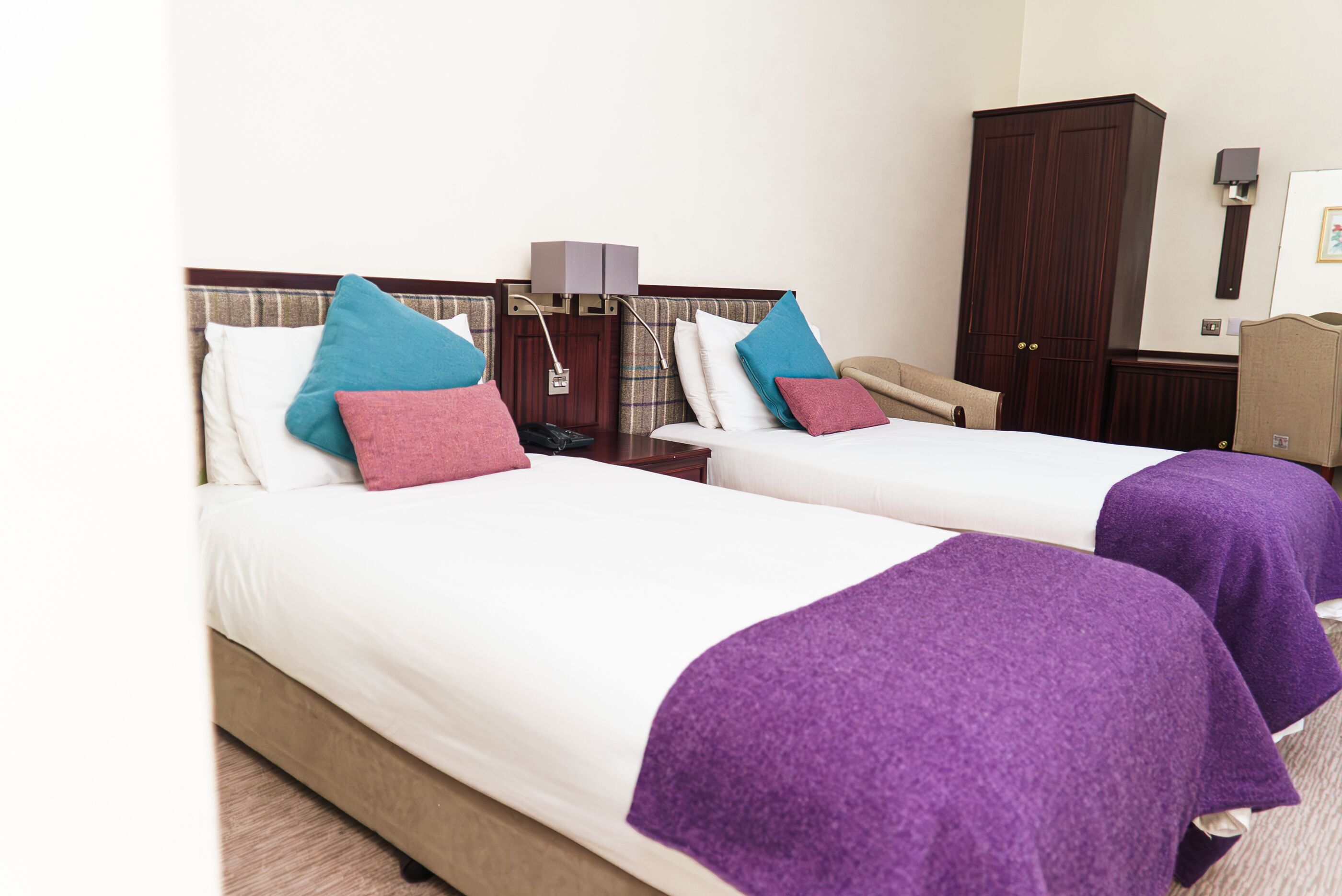 A hotel room with two beds, each with a purple blanket and pillows in various colors. There is a wooden wardrobe and a desk with a telephone.