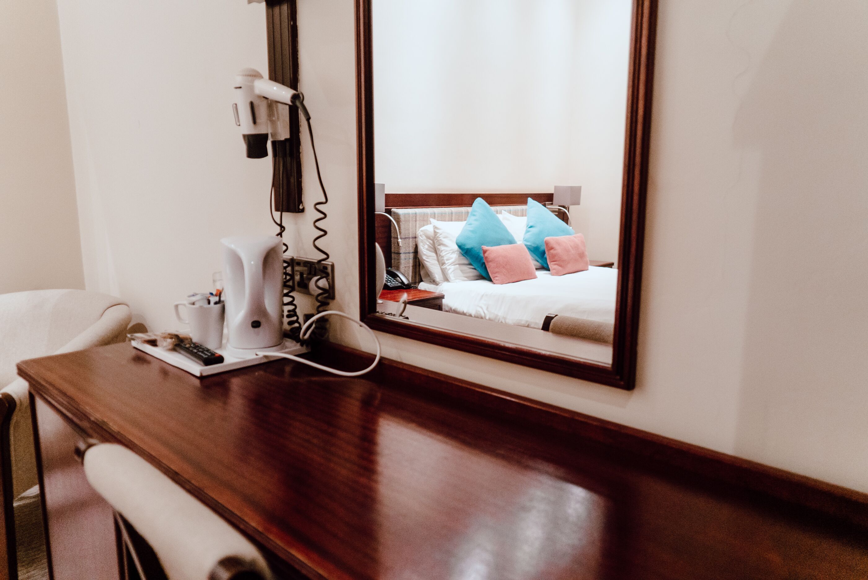 Hotelkamer met een houten bureau, een haardroger, een telefoon en een spiegel die het bed met kussens weerspiegelt.