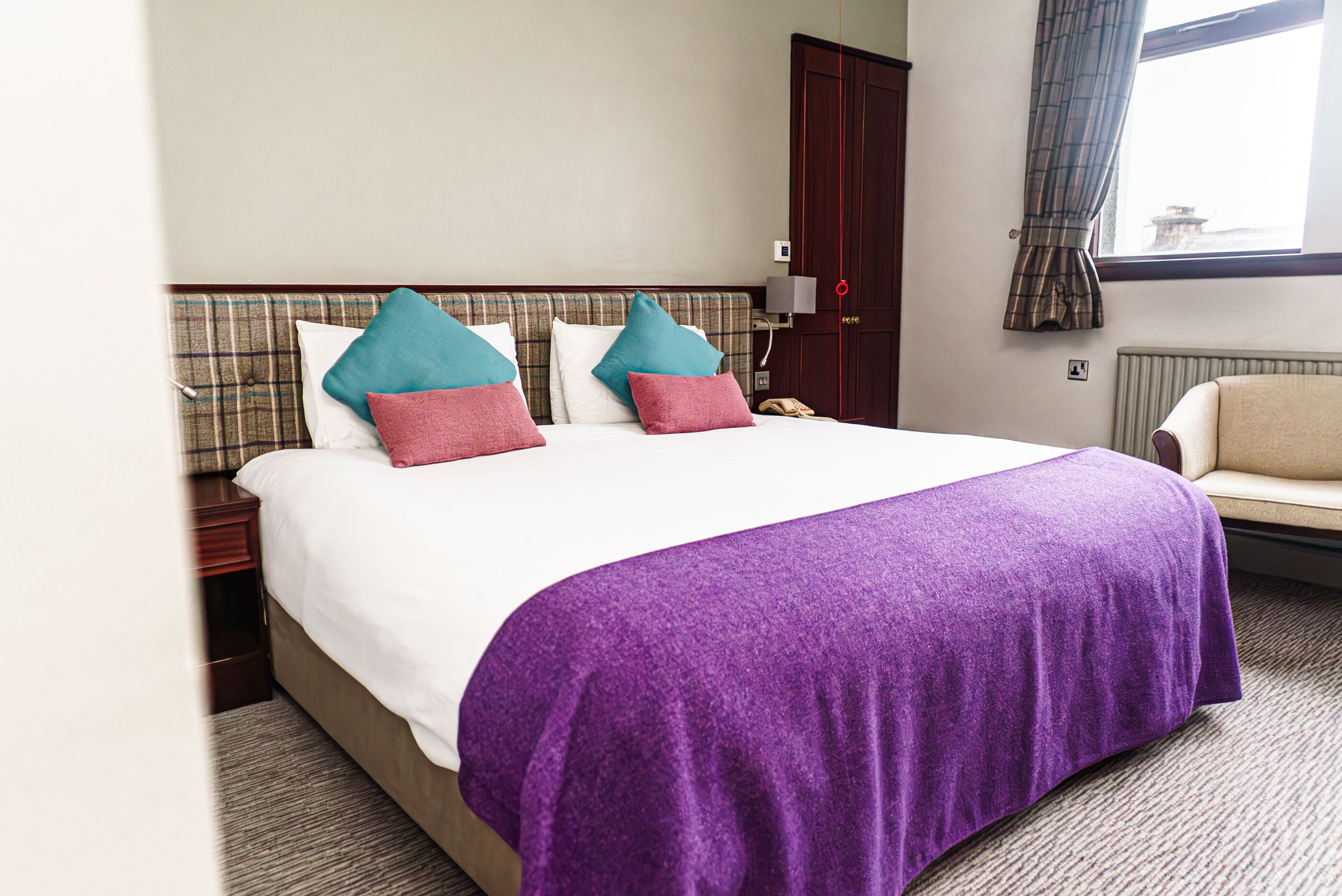 A hotel room with a large bed, two pillows, a nightstand, a chair, a window with curtains, and a door.