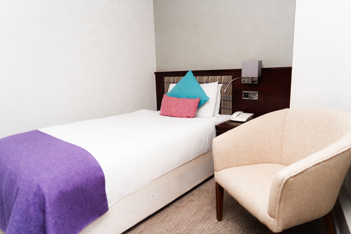Una habitación de hotel con una cama, una silla, una mesita de noche y una lámpara de pared.