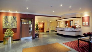 A hotel lobby with a reception desk, a large abstract painting, a potted plant, and a red carpeted area.
