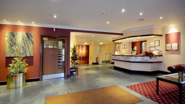 A hotel lobby with a reception desk, a large abstract painting, a potted plant, and a red carpeted area. - Caladh Inn (Stornoway)