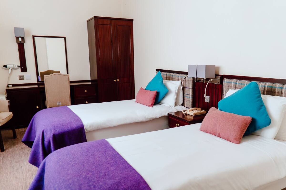 Habitación de hotel con dos camas, un armario, un escritorio con silla y una mesita de noche con lámpara.