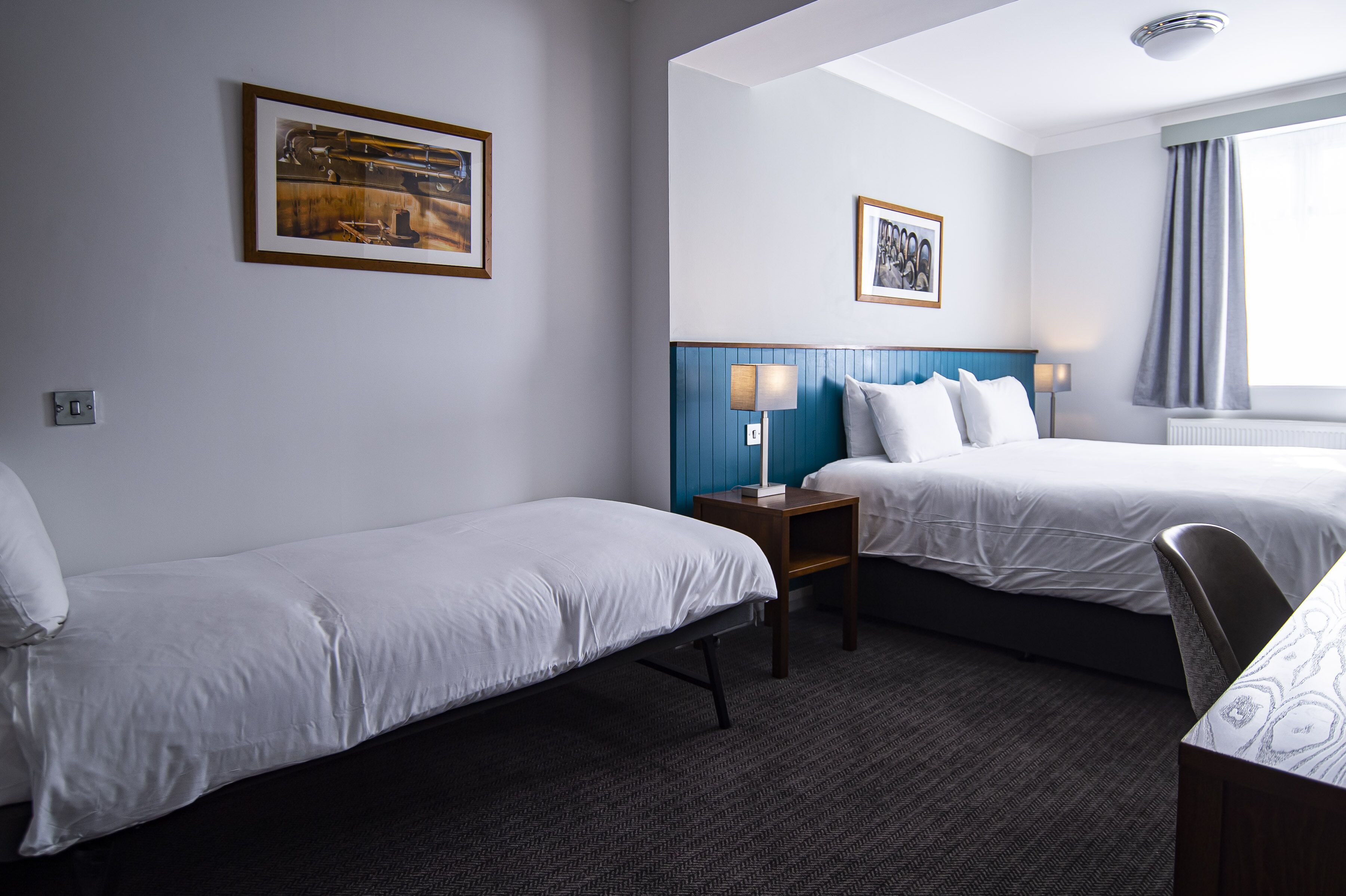 Habitación de hotel con dos camas individuales, una mesita pequeña, dos mesitas de noche con lámparas y cuadros enmarcados en las paredes.